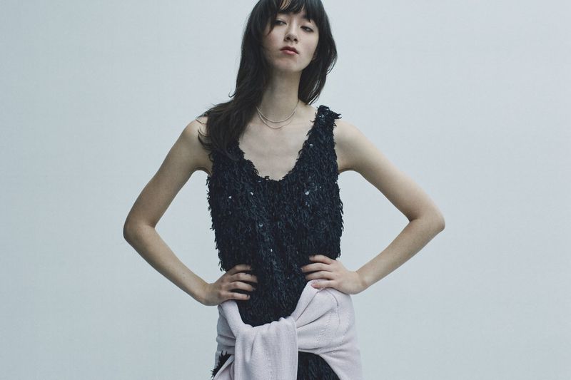 Model wearing a black textured sleeveless top with a light pink sweater tied around her waist against a grey background.