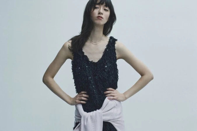 Model wearing a black textured sleeveless top with a light pink sweater tied around her waist against a grey background.