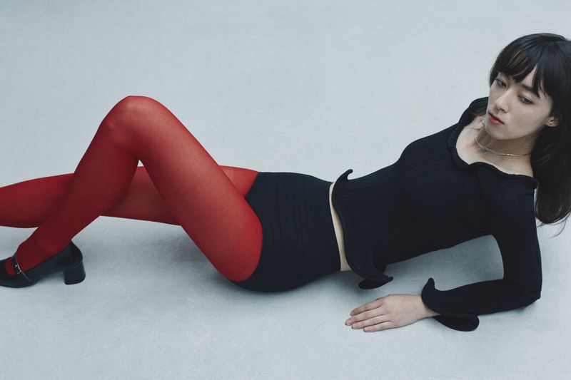 A woman poses in a studio wearing a long-sleeved black top, red tights, black shoes, and black bottoms against a neutral backdrop.