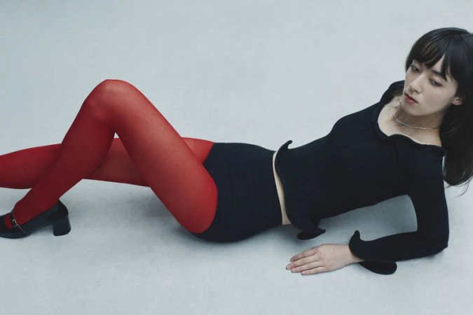 A woman poses in a studio wearing a long-sleeved black top, red tights, black shoes, and black bottoms against a neutral backdrop.
