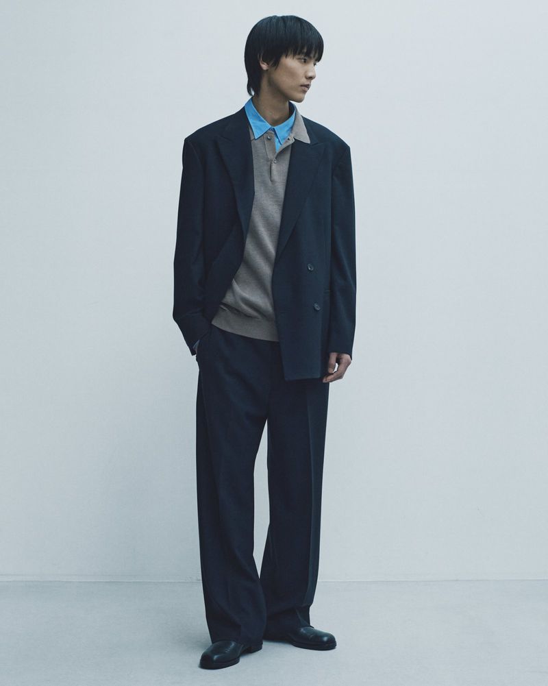 Young man in oversized navy suit, standing against pale backdrop, exudes calm and modern style with neutral knit polo shirt.