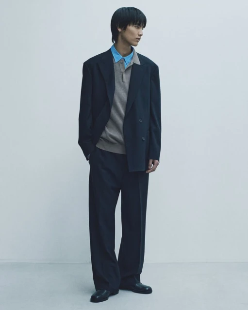 Young man in oversized navy suit, standing against pale backdrop, exudes calm and modern style with neutral knit polo shirt.