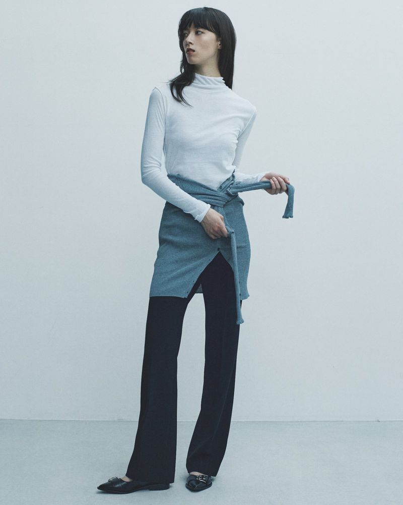 A woman wearing a white long-sleeved shirt, black pants, and a blue-grey wrap is standing against a white background in a fashion shot.