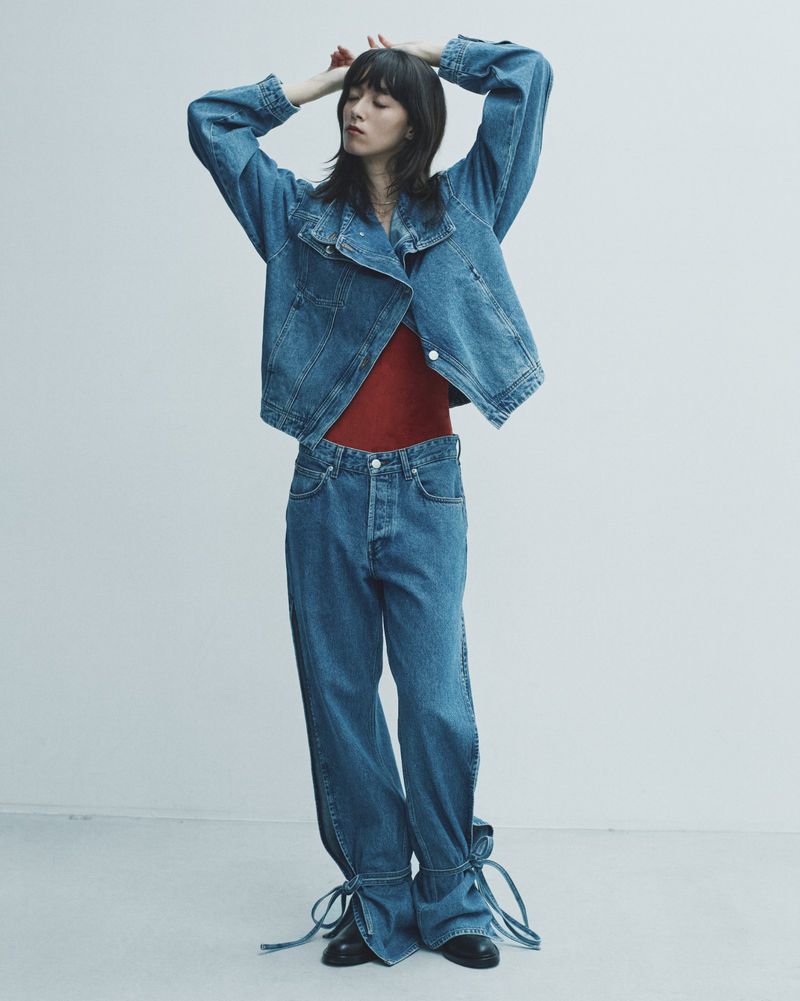 A woman stands in a photo studio wearing a denim jacket, red shirt, and denim pants with ties at the ankle in a full shot.