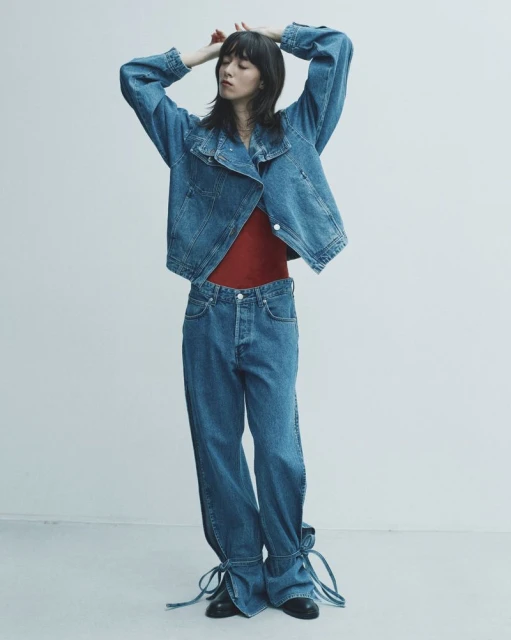 A woman stands in a photo studio wearing a denim jacket, red shirt, and denim pants with ties at the ankle in a full shot.