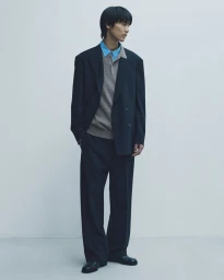 A male model wearing a dark oversized suit jacket over a beige knit polo shirt against a plain grey studio backdrop.