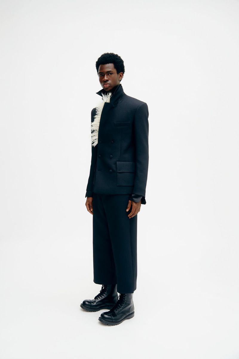 Full-length studio shot of an adult male in a black suit, black boots, and a feathered accessory against a bright white background.