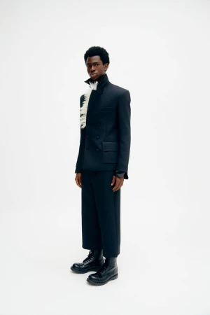 Full-length studio shot of an adult male in a black suit, black boots, and a feathered accessory against a bright white background.