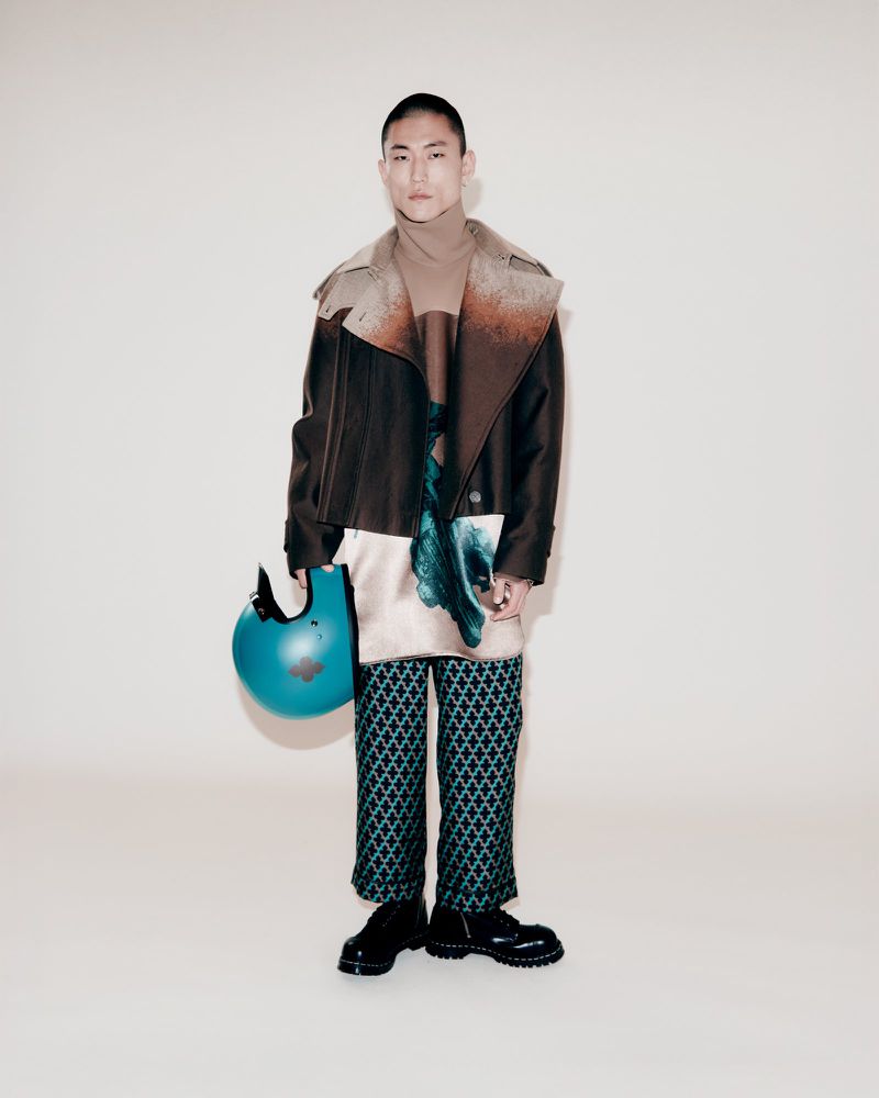 Asian man in patterned pants, jacket, and boots poses indoors, holding a helmet against a neutral backdrop in a studio shot.