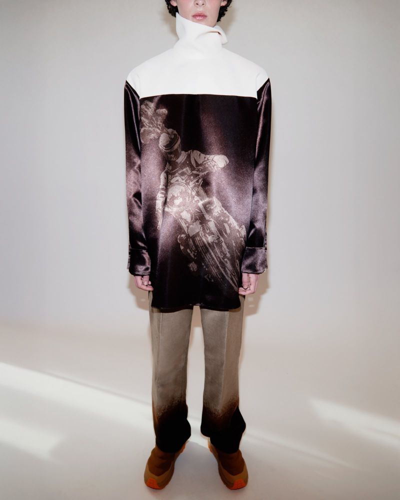 Model wearing a white and dark satin top with graphic print and neutral trousers against a plain seamless white backdrop.