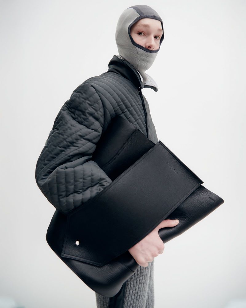 Fashionable person wearing a grey balaclava and black jacket holding a large black leather envelope bag in a bright studio.