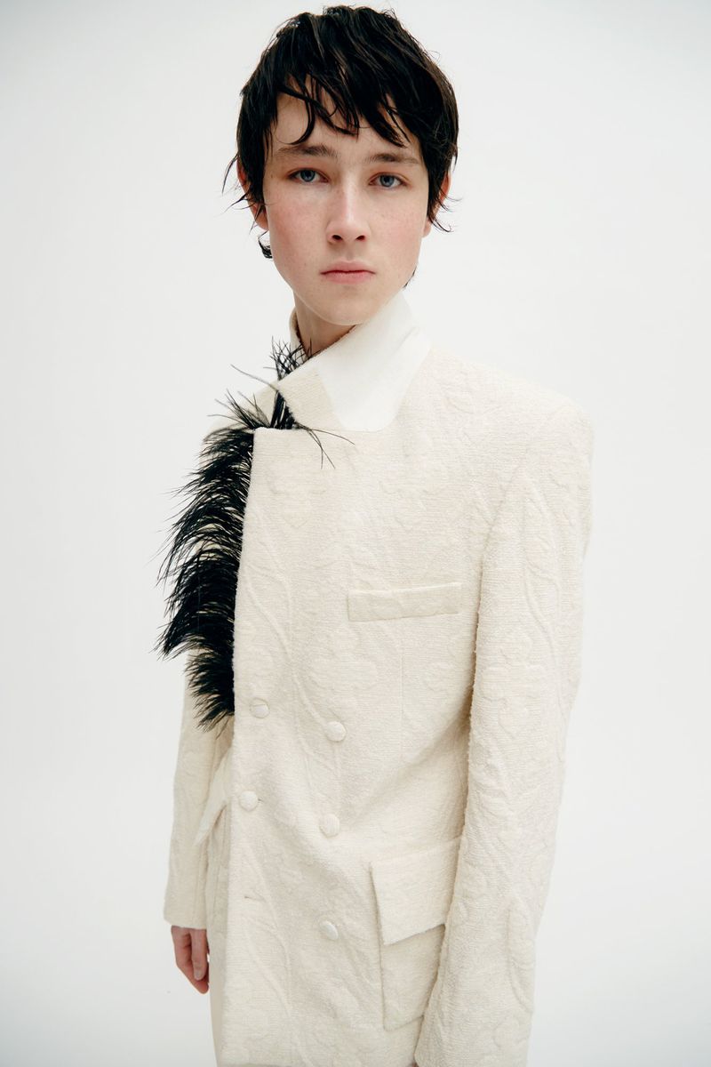 Portrait of a young adult in a patterned white suit with black feathers, posing in a bright photo studio against a white backdrop.