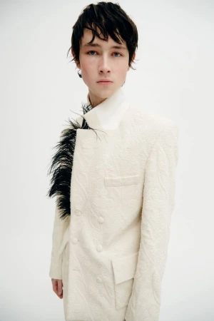 Portrait of a young adult in a patterned white suit with black feathers, posing in a bright photo studio against a white backdrop.