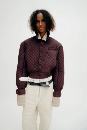 Woman in a quilted bomber jacket, ribbed pants, and belts against a white backdrop in a fashion-forward studio portrait. 