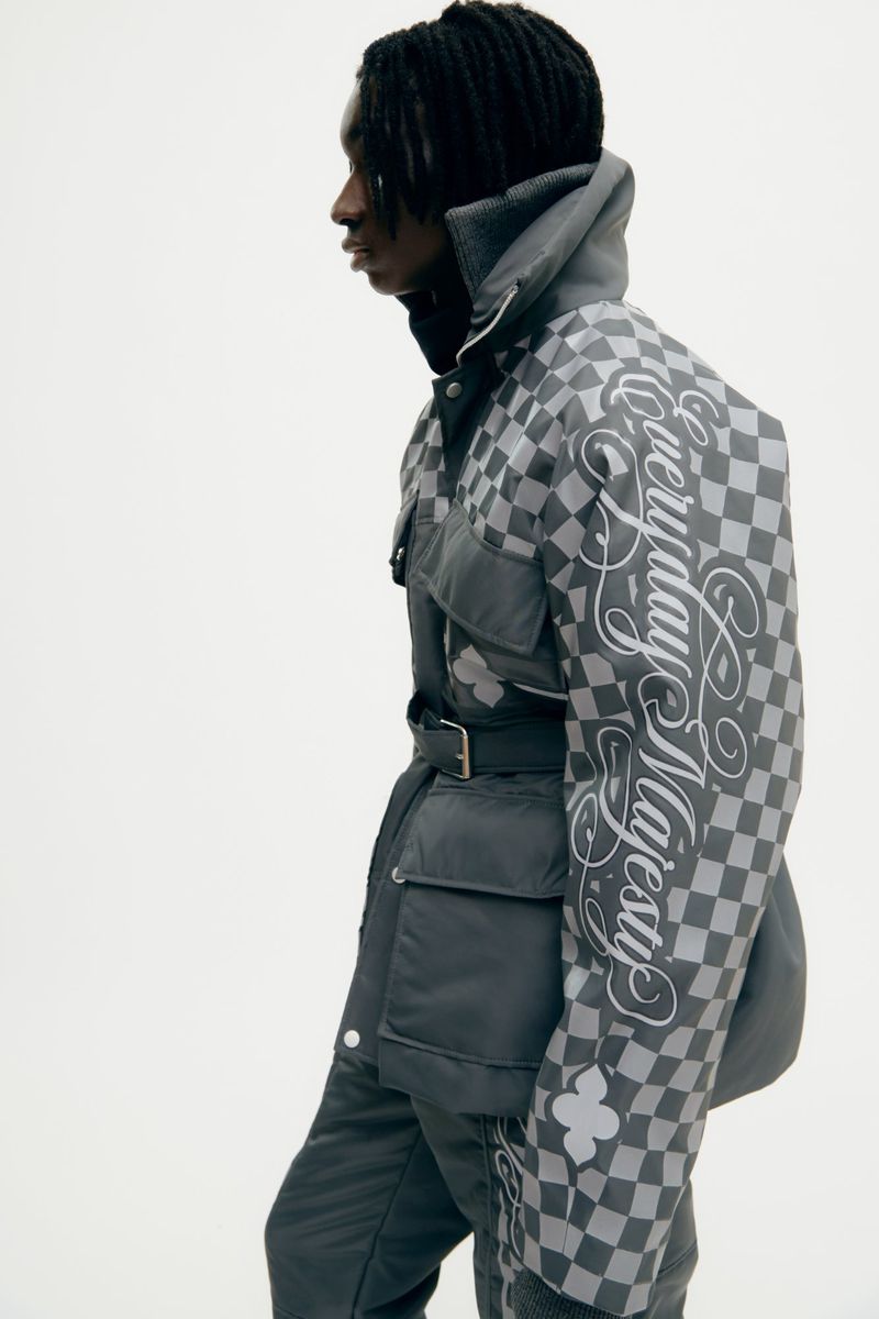 A slender young Black male model poses sideways in a grey check-patterned jacket with cursive text, belted at the waist in a studio shot.