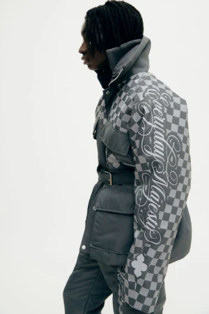 A slender young Black male model poses sideways in a grey check-patterned jacket with cursive text, belted at the waist in a studio shot.