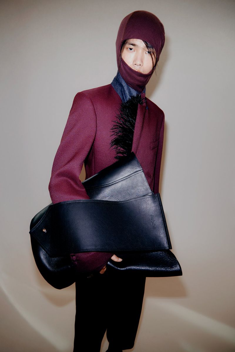 Asian male model in burgundy suit and balaclava holds black leather bag in indoor studio fashion shot. Modern, sleek, and stylish.