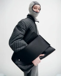 Model wearing a grey balaclava and quilted jacket holding a large black leather bag against a white seamless background