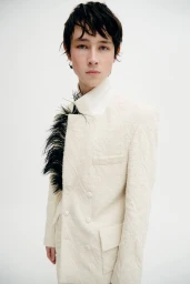 Model wearing a cream patterned double-breasted jacket accented with black feathers against a seamless white background.
