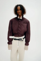 Model wearing a quilted burgundy jacket and white textured trousers against a seamless white background.