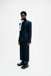 Model wearing a black structured jacket and trousers with white ruffles against a white backdrop for Denzil Patrick AW25.