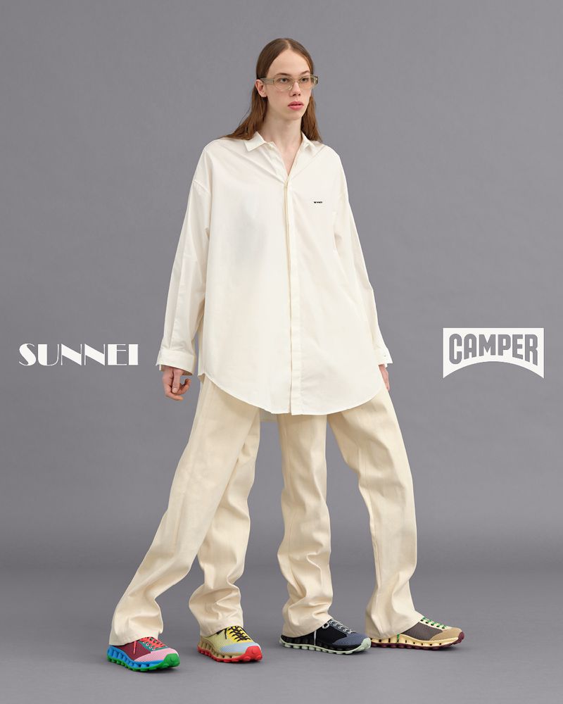 Fashion editorial shot of a tall, slender model posing in a white oversized shirt, beige trousers and colorful sneakers against a grey background.