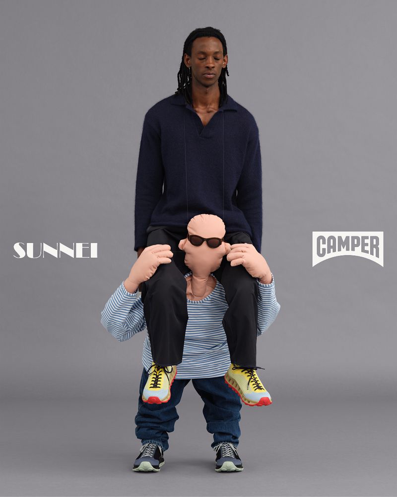 Two men in an indoor studio setting, one carrying the other on his shoulders, showcasing fashion clothing with brand logos. 