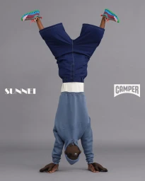 Model doing a handstand wearing a blue hoodie, blue shorts, and colourful sneakers against a grey studio backdrop.