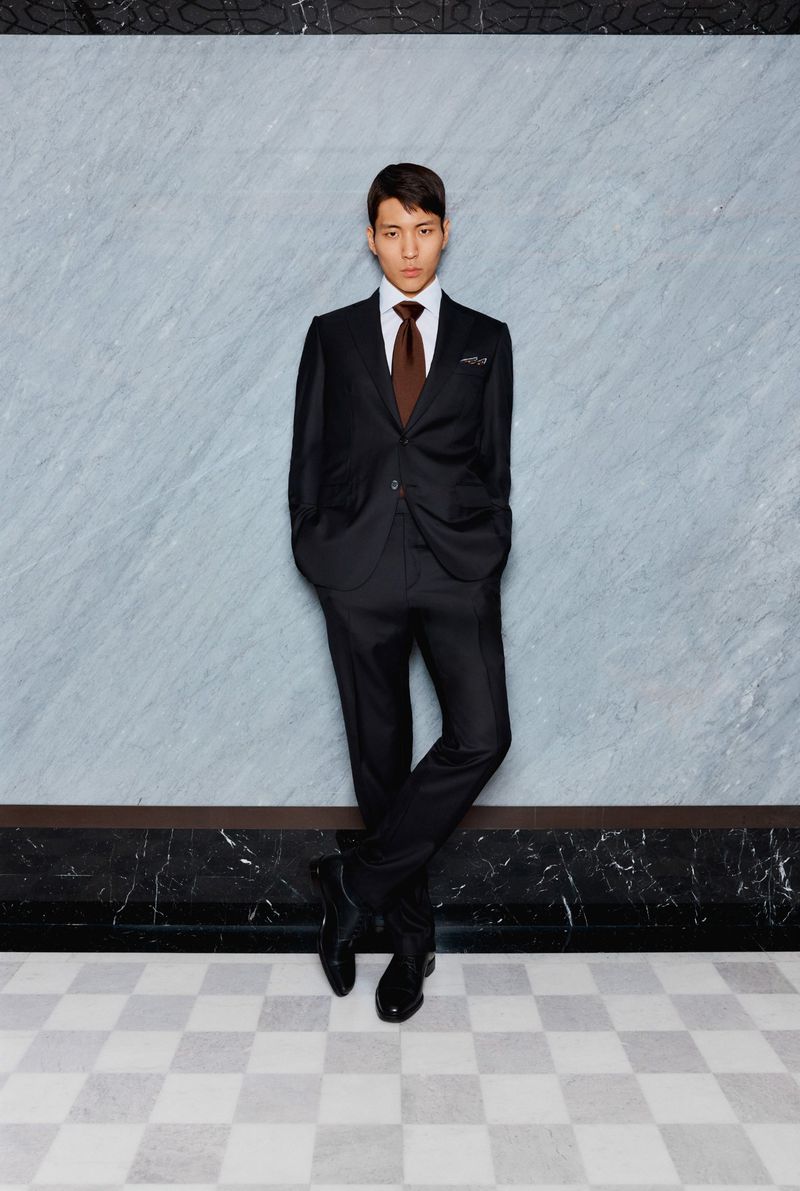Elegant Asian man in a black suit with brown tie poses confidently against a marble wall and checkered floor, demonstrating formal style.
