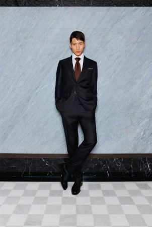 Elegant Asian man in a black suit with brown tie poses confidently against a marble wall and checkered floor, demonstrating formal style.