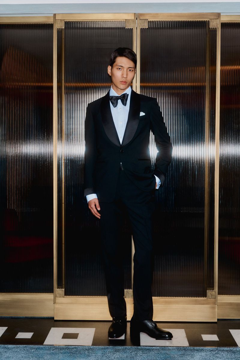 Full-length portrait of an Asian man in a black tuxedo with bow tie, posing in front of a golden textured door and patterned floor.