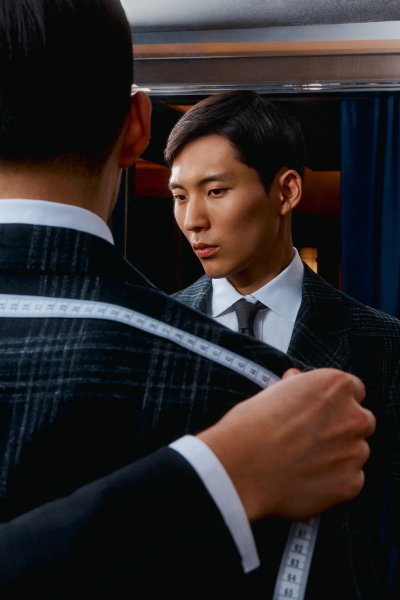 Young Asian man in a tailored suit being measured with a tape measure by a tailor in an indoor fitting at a designer store.
