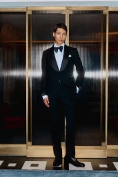 Man wearing a black tuxedo with a bow tie standing before ribbed glass doors with brass frames.