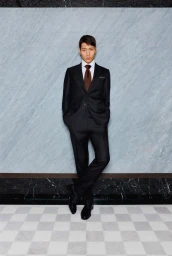Man wearing a black suit and brown tie leaning against a blue marble wall for Harrods Tailoring 2025 editorial