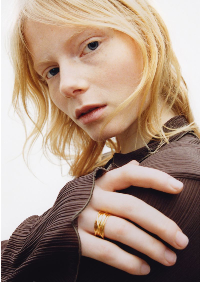 Close-up of a fair-skinned adult woman wearing a brown shirt and three gold rings on her finger, posing in an indoor setting.