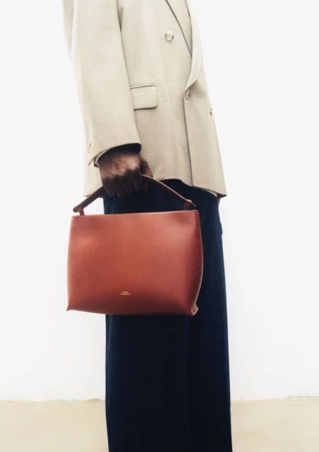 Person holds a brown leather tote bag. The subject wears a beige blazer and a long navy skirt against a white background. Minimal style.