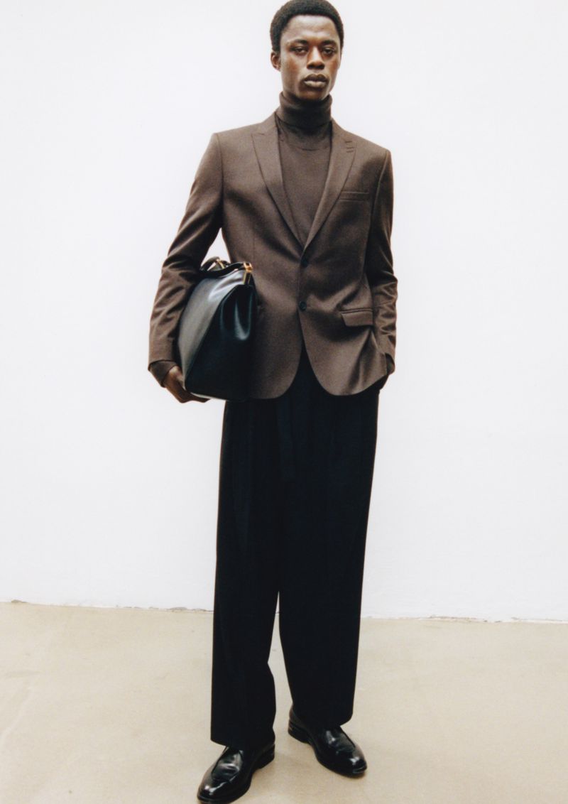 Man poses in blazer and trousers, holding large black bag against a white backdrop in a fashion editorial shot in the studio.