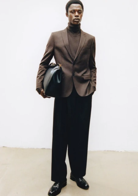 Man poses in blazer and trousers, holding large black bag against a white backdrop in a fashion editorial shot in the studio.