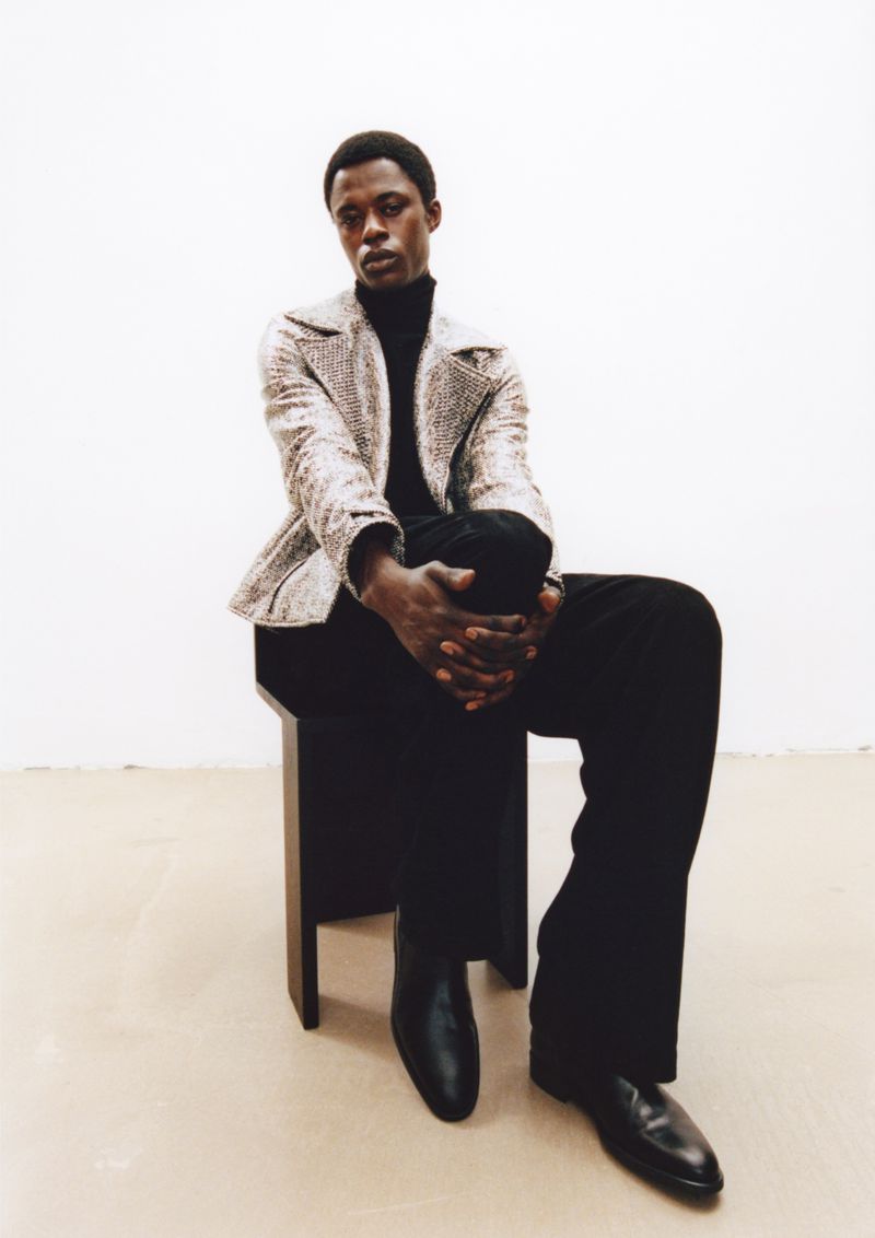 Black man seated on a stool wearing a patterned jacket, black turtleneck, and black trousers against a neutral background.