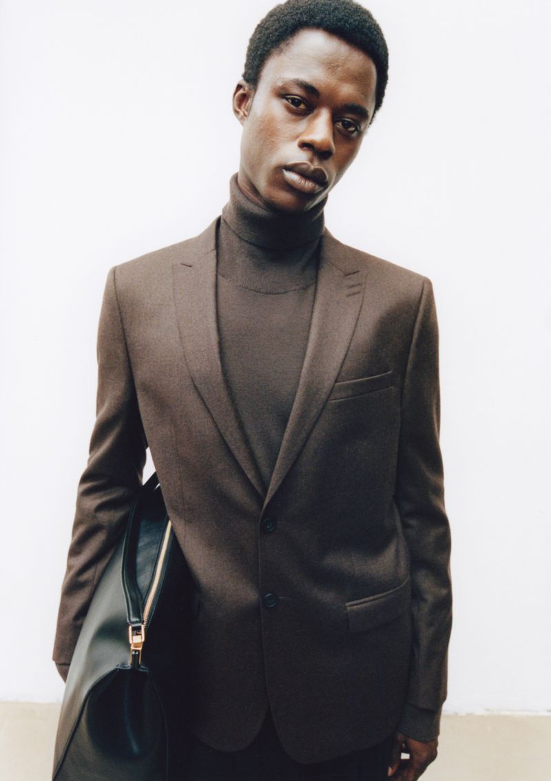 Portrait of Black male model in brown suit and turtleneck against a white wall, accessorized with a sleek black leather bag.