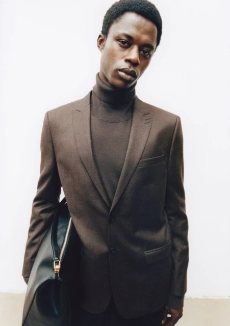 Portrait of Black male model in brown suit and turtleneck against a white wall, accessorized with a sleek black leather bag.