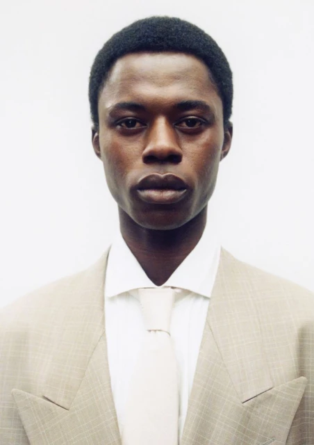 Portrait of a stylish African American man in a beige suit, white dress shirt, and tie, set against a plain white backdrop.