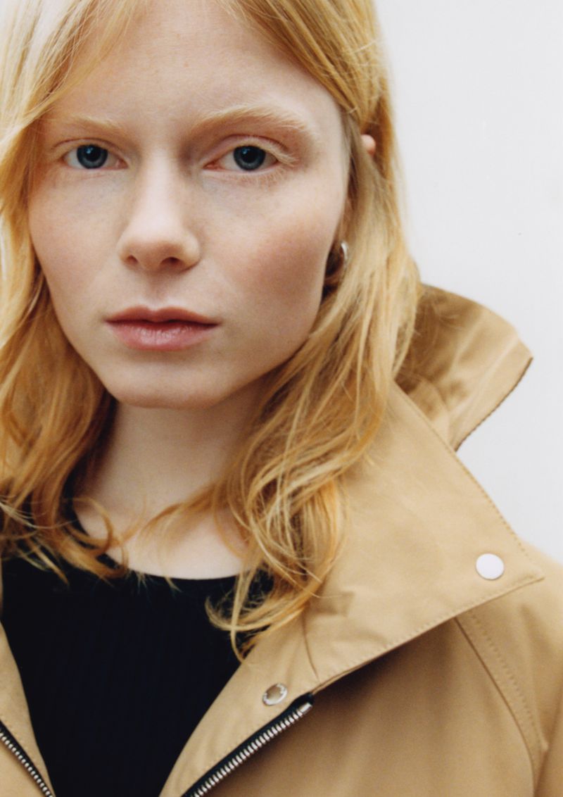 Close-up portrait of a female model with strawberry blonde hair wearing a tan hooded jacket against a white background