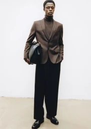 Black man wearing a brown blazer, turtleneck, and black trousers holding a dark leather bag against a white seamless background