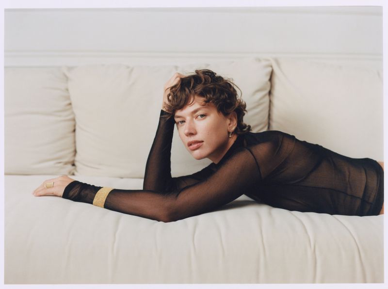 Brunette woman in a black mesh top lies on a white cushioned surface with one hand on her head and looks directly at the camera.