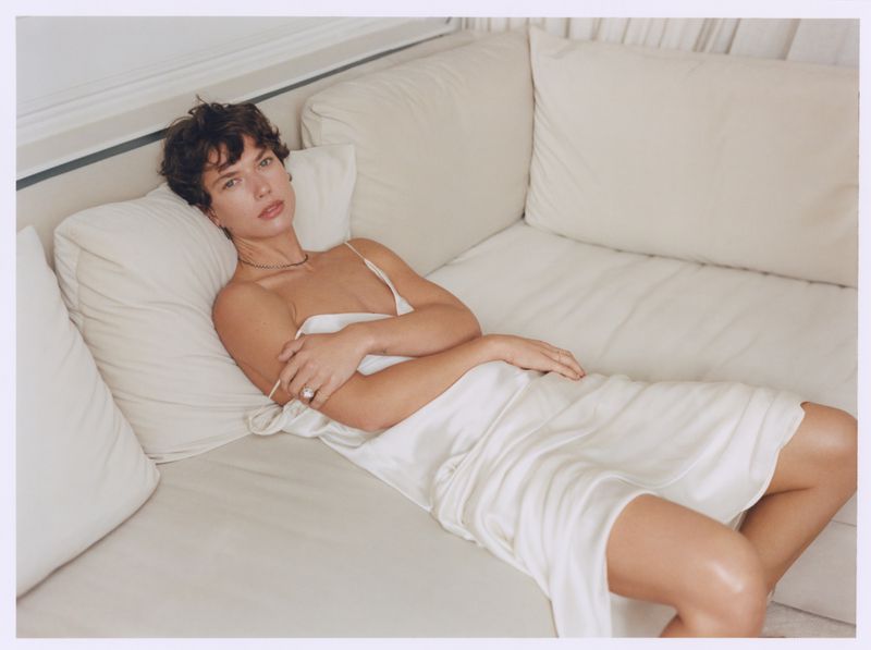 Woman lying on a light beige couch. She has short brown hair and is partially covered with a white sheet or dress.