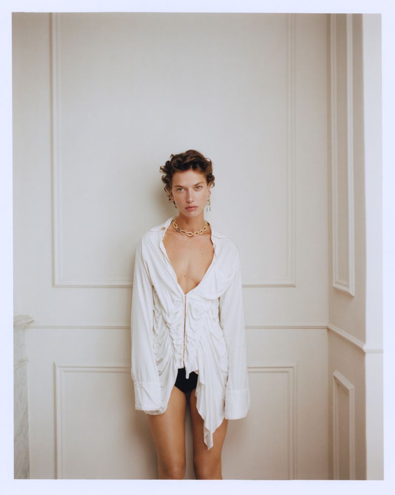 A fashionable adult female poses indoors wearing a white collared shirt, gold necklace, and black bottoms in a medium shot.