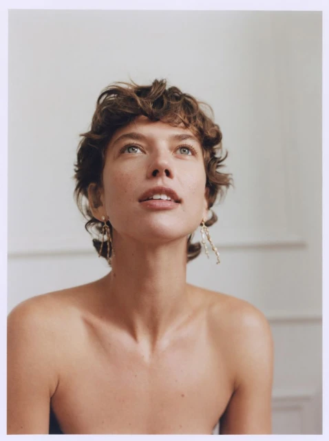 Portrait of a young adult woman with curly brown hair looking up with a neutral expression, wearing dangling gold earrings against a white wall.