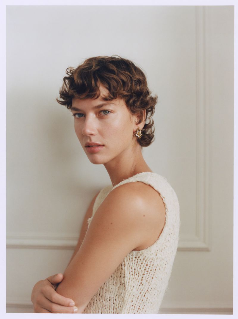 Portrait of a woman with short brown hair wearing a cream-colored knit top, posing against a white wall in an indoor setting.