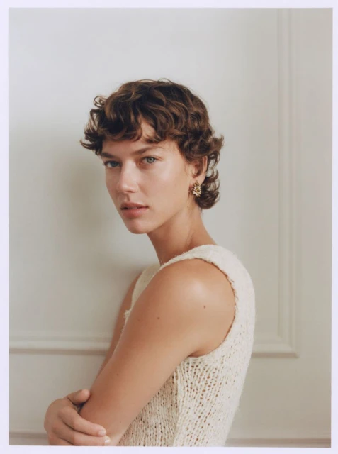 Portrait of a woman with short brown hair wearing a cream-colored knit top, posing against a white wall in an indoor setting.